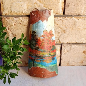 May include: A curved terracotta roof tile featuring a hand-painted landscape scene of a forest with orange and green trees, a blue sky, and a reflective body of water. The tile has a rustic brown border and includes a thin twine loop for hanging.
