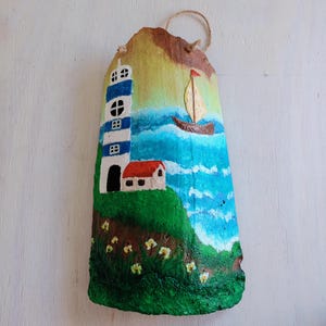 May include: Hand-painted coconut shell with a seascape scene. The scene features a blue and white lighthouse, a red-roofed house, a sailboat on blue water, and green grass with yellow flowers.