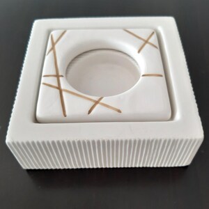 May include: White ceramic candle holder with a square base and a smaller square insert with a circular opening. The insert has a gold geometric pattern.