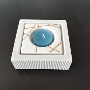 May include: White ceramic candle holder with a square shape and a raised, textured rim. The holder has a circular cutout in the center with a blue teal candle inside. The holder is decorated with gold lines.