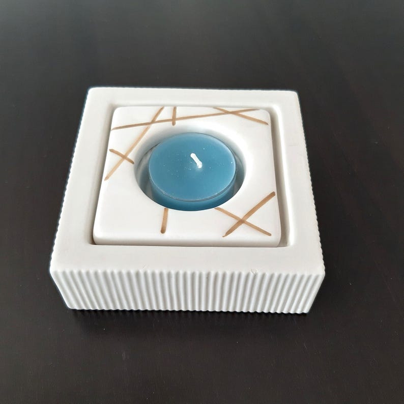 May include: White ceramic candle holder with a square shape and a raised, textured rim. The holder has a circular cutout in the center with a blue teal candle inside. The holder is decorated with gold lines.