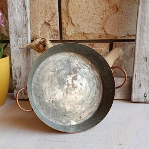 May include: A round, shallow metal pan with a weathered, silver-toned interior surface showing signs of wear and oxidation. The pan features two small, curved copper-coloured handles attached to opposite sides with visible rivets on the rim.