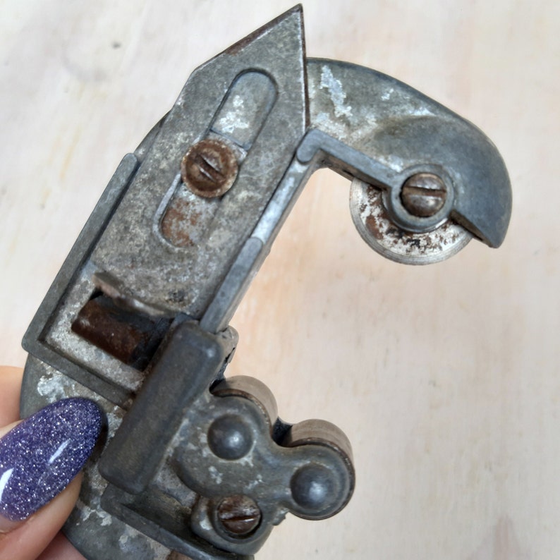 May include: A vintage metal pipe cutter with a silver finish and a black handle. The cutter has a rotating wheel and a lever for cutting pipes.