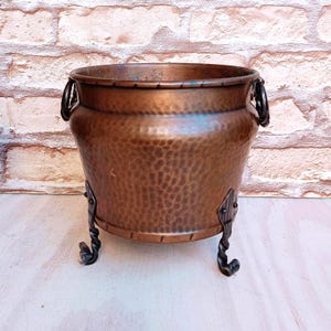May include: A round, hammered copper planter with a wide rim and a dark, weathered patina. The vessel features two side ring handles and is supported by three ornate, twisted black metal legs that curve outward at the base for stability.