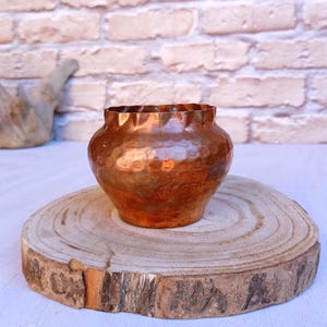 May include: A copper-toned vase with a textured finish and a decorative, ruffled rim. The vase sits on a round wooden slice, highlighting its rustic appeal. The background features a brick wall.