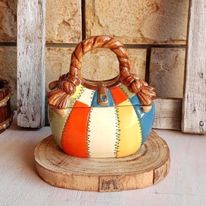 May include: A ceramic handbag-shaped container featuring a patchwork design with panels of orange, cream, yellow, and blue. The piece has a brown rope-textured handle and a central clasp detail with a small blue accent, finished with black stitching.
