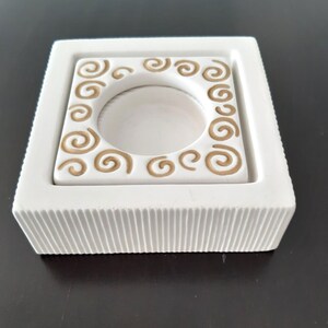 May include: White ceramic candle holder with a square base and a raised inner square with a circular cutout. The inner square has a decorative border of gold swirls.