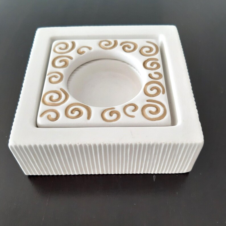 May include: White ceramic candle holder with a square base and a raised inner square with a circular cutout. The inner square has a decorative border of gold swirls.