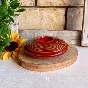 Può includere: Un portacandele in ceramica circolare e basso, caratterizzato da una finitura lucida rosso intenso e linee concentriche color marrone dorato a spirale che si irradiano dall'apertura centrale. L'oggetto è posizionato su una fetta di legno.