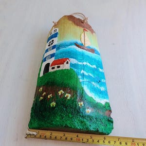 May include: Hand-painted wooden wall hanging featuring a lighthouse, a sailboat, and a small house on a green hill with yellow flowers. The background is a blue ocean with white clouds.