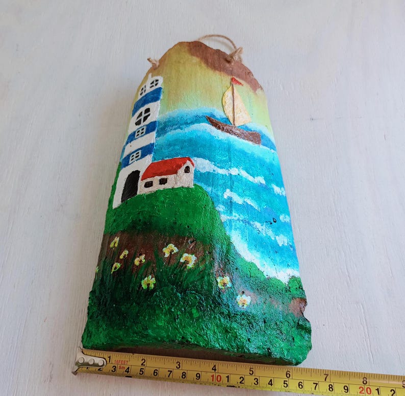 May include: Hand-painted wooden wall hanging featuring a lighthouse, a sailboat, and a small house on a green hill with yellow flowers. The background is a blue ocean with white clouds.