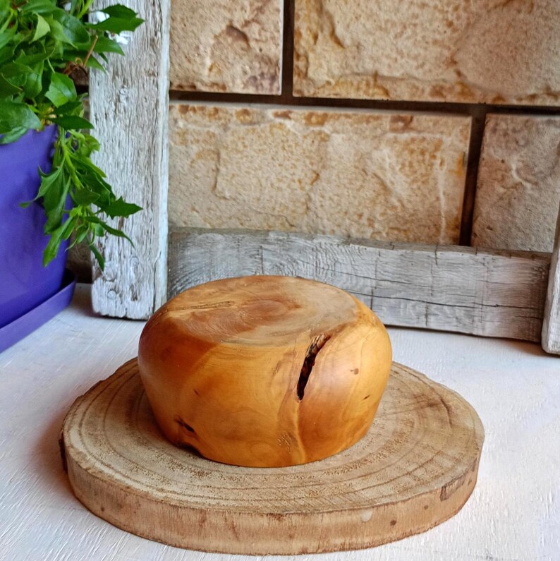 May include: A light brown wooden bowl with a smooth, rounded exterior and a visible natural crack on the side. The bowl is positioned on top of a flat, circular wooden slice base that features visible tree rings and a rough, textured bark edge.