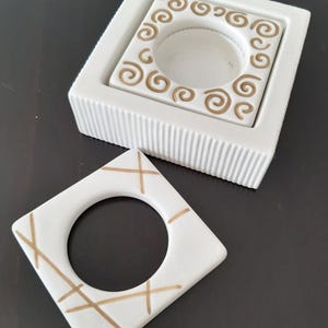 May include: Two white ceramic candle holders with gold designs. One holder has a swirl design and the other has a criss-cross pattern.