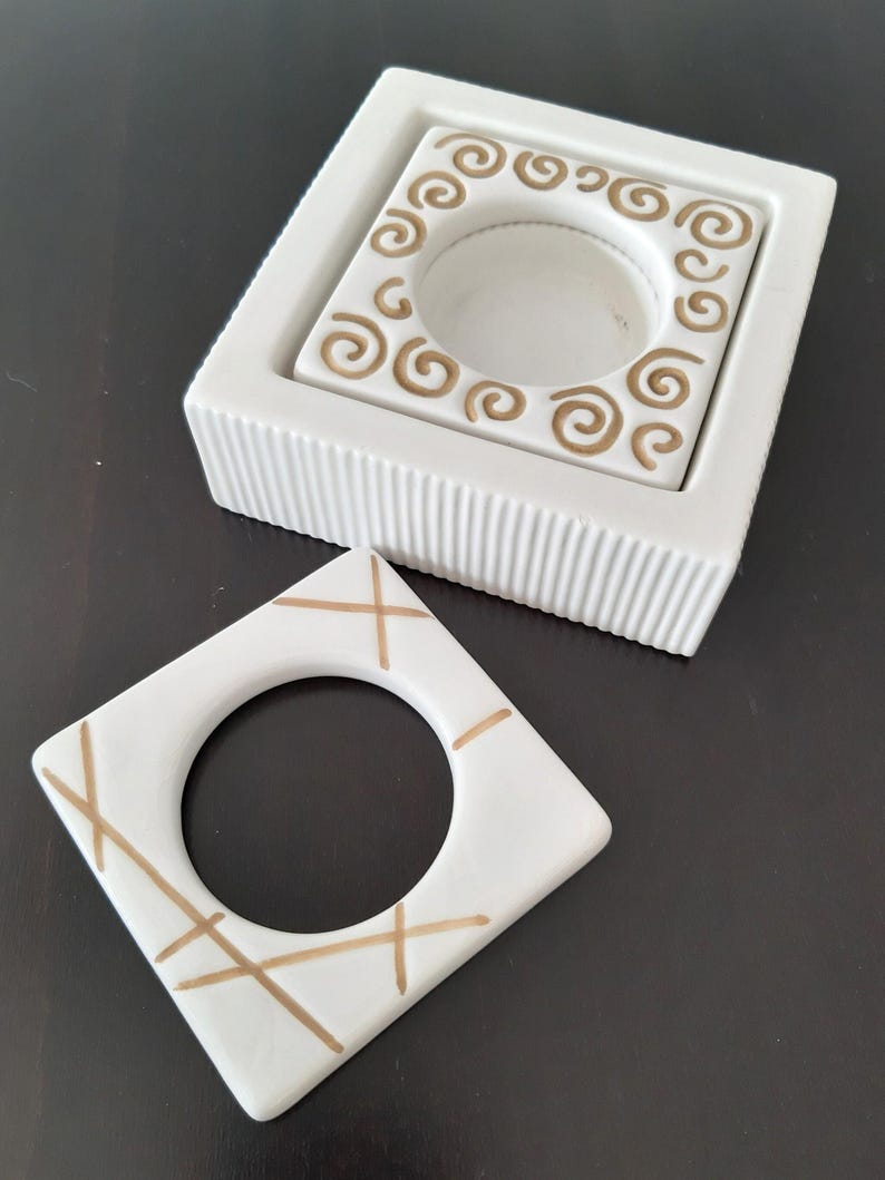May include: Two white ceramic candle holders with gold designs. One holder has a swirl design and the other has a criss-cross pattern.