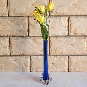 May include: A tall, cobalt blue glass vase with a flared base holds several yellow flowers with green stems and leaves. The vase has a slender, elongated shape. The background is a light brown brick wall.