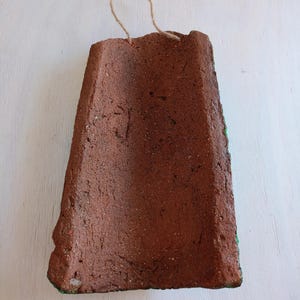 May include: A terracotta-colored clay tile with a rough texture and a green edge. The tile has a string attached to it, suggesting it is a hanging ornament.