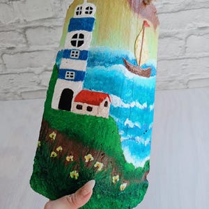 May include: A hand-painted seascape scene on a piece of wood. The scene features a lighthouse with blue and white stripes, a small house with a red roof, a sailboat on the water, and yellow flowers.