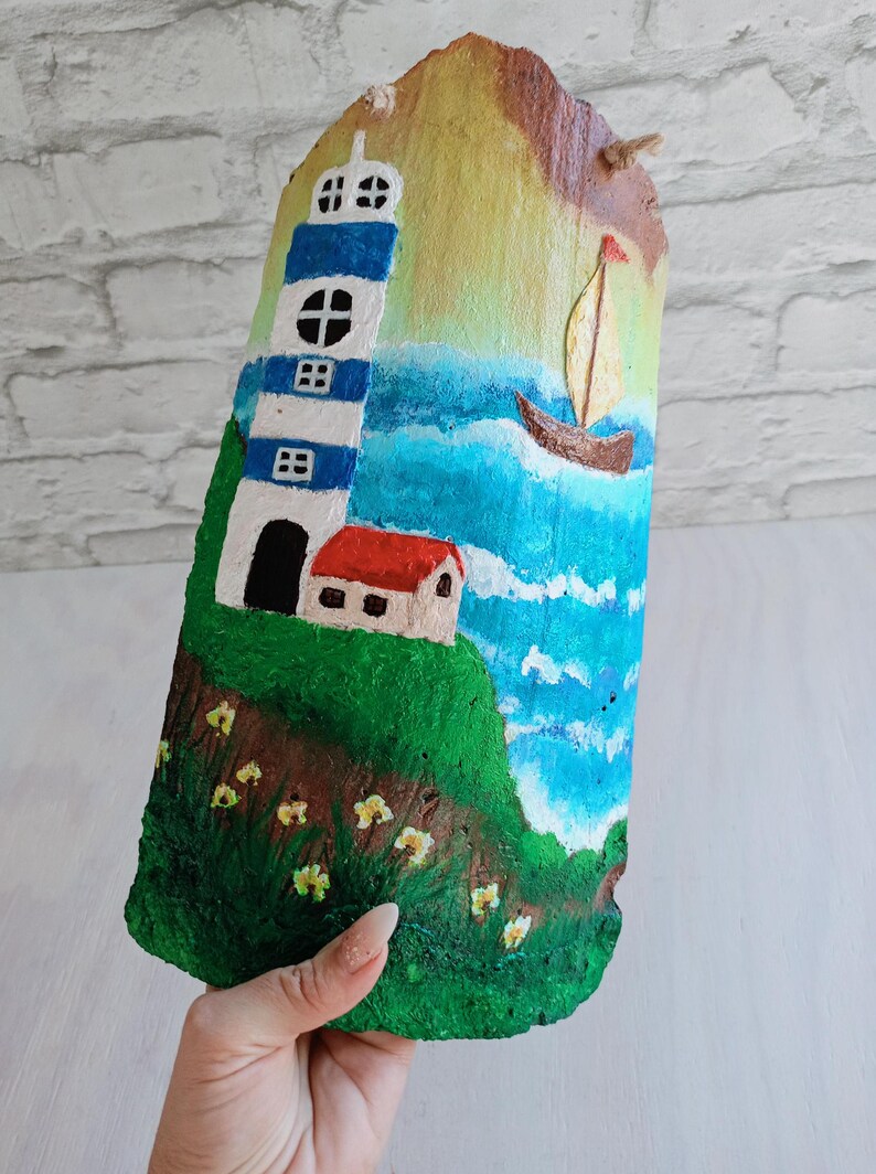 May include: A hand-painted seascape scene on a piece of wood. The scene features a lighthouse with blue and white stripes, a small house with a red roof, a sailboat on the water, and yellow flowers.