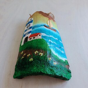 May include: Hand-painted wooden seascape with a lighthouse, a small house, a sailboat, and green grass with yellow flowers. The background is a blue ocean with white waves.