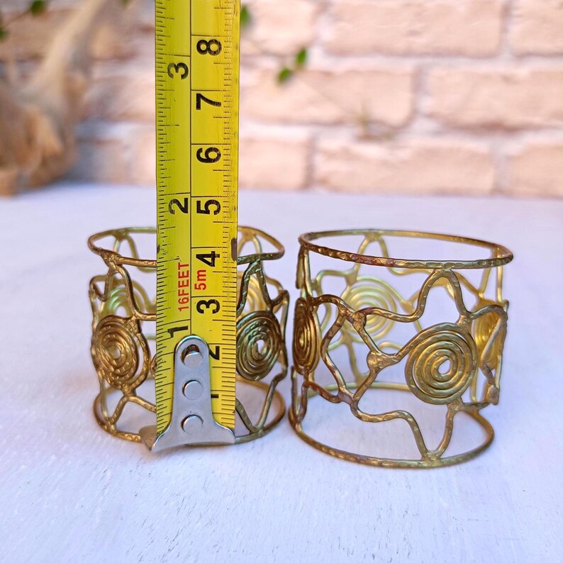 Pu&ograve; includere: Due portatovaglioli in metallo color oro con un design traforato con spirali e motivi a forma di stella. Ogni anello misura circa 5 cm di altezza. Gli anelli sono esposti su una superficie chiara.
