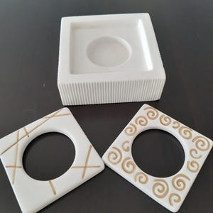 May include: Three white ceramic candle holders with gold accents. The holder on the top has a square base and a round opening. The two holders on the bottom are square with a round opening and different gold designs.