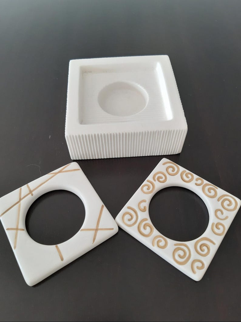 May include: Three white ceramic candle holders with gold accents. The holder on the top has a square base and a round opening. The two holders on the bottom are square with a round opening and different gold designs.