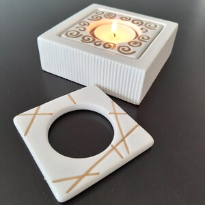 May include: Two white ceramic candle holders with gold accents. The larger holder has a square base and a square top with a circular opening for a tea light candle. The smaller holder is a square with a circular opening in the center and gold lines across the surface.