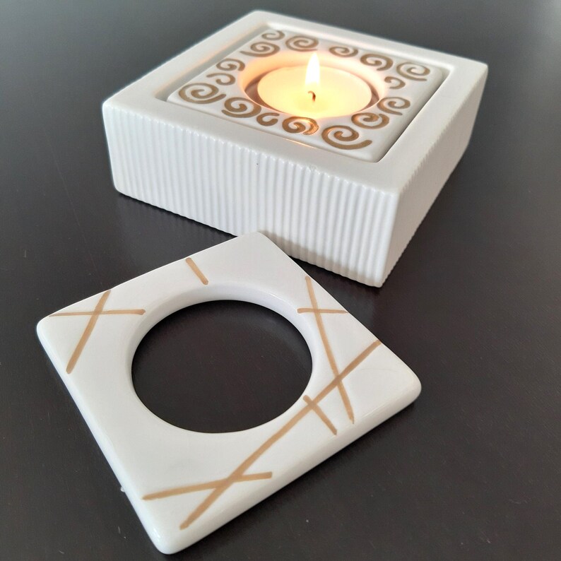 May include: Two white ceramic candle holders with gold accents. The larger holder has a square base and a square top with a circular opening for a tea light candle. The smaller holder is a square with a circular opening in the center and gold lines across the surface.