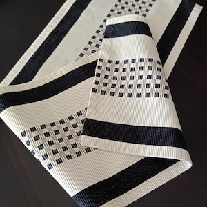 May include: Cream-colored placemat and napkin set with black and tan geometric patterns. The placemat has a striped border and a square pattern in the center. The napkin is folded and features a similar design. The set is made of a textured fabric.