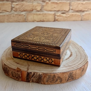 May include: A small wooden box with intricate marquetry designs in shades of brown and gold. The box is closed and sitting on a wooden surface.