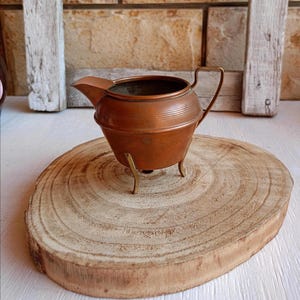 May include: A copper creamer pitcher featuring a rounded body with horizontal ribbed detailing, a small pouring spout, a thin curved handle, and three slender brass-toned metal legs that elevate the vessel from the surface.