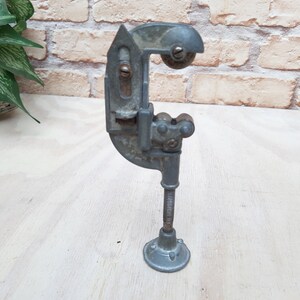 May include: A silver pipe cutter with a C-shaped clamp and a rotating wheel. The cutter has a base with a threaded rod and a foot pedal.