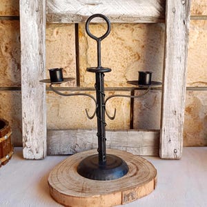 May include: A black metal candle holder with two arms, each holding a small candle cup. The holder has a circular base and a decorative, twisted central stem. It is displayed on a wooden slice.