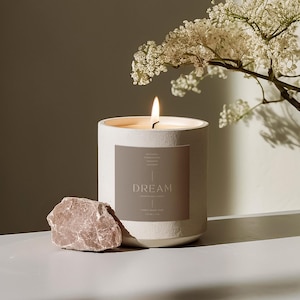 Op de afbeelding: Een witte kaars met een bruin label waarop in een minimalistisch lettertype "DREAM" staat. De kaars is aangestoken en de vlam is zichtbaar. De kaars staat op een wit oppervlak met een kleine roze steen ernaast. Op de achtergrond is een witte bloemen tak te zien.