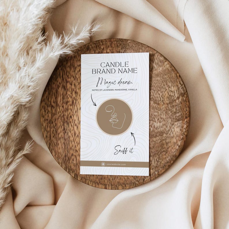 May include: A white card with brown text and a brown circle with a line drawing of a face. The text reads "CANDLE BRAND NAME Magic dream NOTES OF LAVENDER, MANDARINE, VANILLA Sniff it yourwebsite.com"