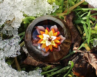 Puede incluir: Un pisapapeles de vidrio con un diseño de flor colorido. La flor tiene pétalos amarillos, naranjas, rojos y blancos. El pisapapeles está sobre un lecho de musgo verde y nieve.