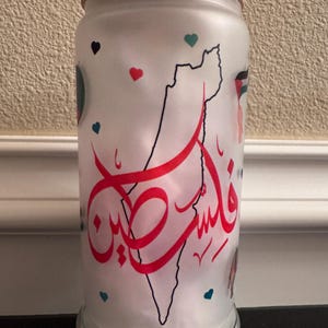 Może przedstawiać: Szklany kubek z matowego szkła z drewnianą pokrywką i przezroczystą słomką. Kubek posiada czarny kontur mapy, czerwoną arabską kaligrafię oraz małe akcenty w kształcie serca i w kolorze morskim. Projekt jest wyśrodkowany na szkle.