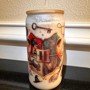May include: A white reusable can with a bamboo lid and a straw. The can has a graphic of the Hogwarts house crests with the Hogwarts school logo at the bottom.