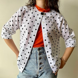 Puede incluir: Una camisa blanca de botones con lunares negros y una camiseta roja debajo. La camisa tiene mangas tres cuartos y cierre de botones.