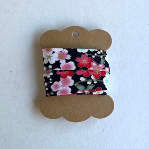 May include: A spool of black fabric ribbon with a floral print of pink and white flowers. The ribbon is on a brown cardboard spool with a scalloped edge.