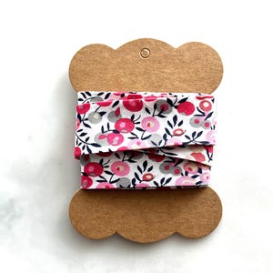 May include: A spool of floral ribbon featuring a repeating pattern of pink flowers, gray accents, and dark green leaves on a white background. The ribbon is wrapped around a brown cardboard spool with a scalloped edge.