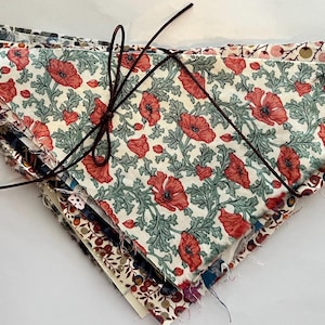 Puede incluir: Un conjunto de cuadrados de tela variados, atados con una cuerda marrón oscuro. La capa superior presenta un estampado floral crema con amapolas rojas y hojas verdes. Otras telas tienen diversos diseños.