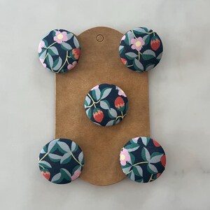 May include: Five circular fabric-covered buttons arranged on a brown cardboard display card. The buttons feature a dark navy blue background with a repeating pattern of light green leaves, small pink flowers, and red strawberry-like fruit shapes.