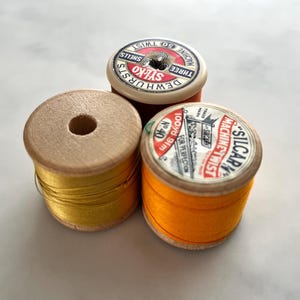Puede incluir: Tres carretes de hilo vintage en tonos dorados, naranja y marrón. Los carretes tienen núcleos de madera y etiquetas de papel con texto. El hilo está enrollado alrededor de los núcleos. Las etiquetas incluyen el texto "SYLKO", "MACHINE TWIST" y "SILCARY".