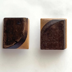 May include: Two rectangular wooden earrings. Each earring is a combination of light and dark brown wood. A dark brown, curved section is on one corner of each earring. The earrings are displayed on a white surface.