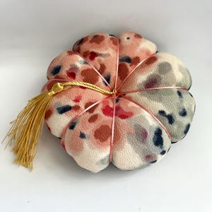 Könnte beinhalten: Ein blütenförmiges Nadelkissen mit einem rosa, roten und blauen gesprenkelten Muster. Es ist mit rosa Band und einer goldenen Quaste gebunden. Der Stoff hat ein strukturiertes Aussehen, und das Design ist dekorativ.