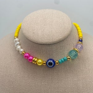 May include: A colorful beaded necklace with a blue evil eye charm. The necklace features yellow, pink, white, and turquoise beads.