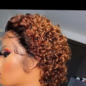 May include: A woman with a curly, two-toned wig. The wig is black at the roots and transitions to a light brown color. The wig is styled in a short, curly bob. The woman is wearing a silver chain necklace.