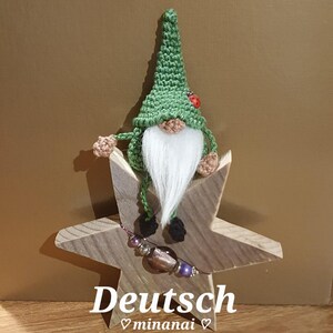 May include: A green crocheted gnome with a white beard sits on a wooden star. The star has a purple and silver beaded bracelet wrapped around it. The gnome has a red ladybug on its hat. The text "Deutsch" and "minanai" is written below the star.