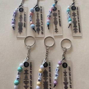May include: Eight clear acrylic keychains with black and white Spotify barcodes and song titles. Each keychain has a small beaded charm with the letters "H" and "S" in different colours.